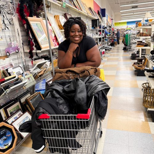 Notre collaboratrice Charmaine souriante dans notre friperie devant un chariot rempli de vêtements comme un manteau en cuir noir